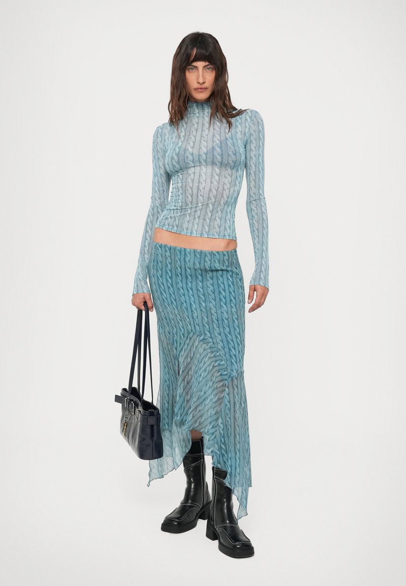 Woman wearing matching blue patterned sheer top and skirt with black chunky boots, holding a black handbag on a white background.