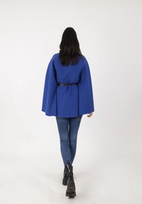 Capa exterior azul de estilo capa con tejido texturizado, ajustada en la cintura con un cinturón negro. Combinada con jeans azules ajustados y botines negros.
