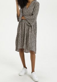 Dunkelgrünes Blumenkleid mit rosa Mustern, V-Ausschnitt und Glockenärmeln; kombiniert mit weißen Sneakern. Weicher Stoff, knielanges Design.