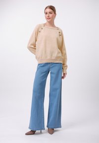Beige strikkegenser med dekorative metallknapper på skuldrene, kombinert med brede, blå jeans og brune spisse hæler.