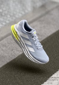 Chaussure de sport gris clair avec un dessus texturé, trois bandes blanches et des accents jaunes vifs sur la semelle, conçue pour la performance et le style.