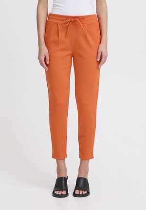Pantalones jogger naranjas con cintura ajustable, pierna tapered y bolsillos laterales, combinados con calzado negro de punta abierta. Textura de tela suave.