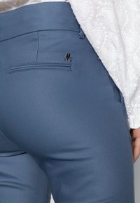 Blå, tettsittende bukser med en baklomme som har en liten svart logo og et glatt, teksturert stoff. Akkompagnert av en hvit mønstret bluse.