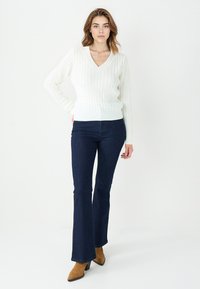 White cable-knit sweater with a V-neck, paired with dark blue flared jeans and brown heeled boots. Smooth textures and fitted design.