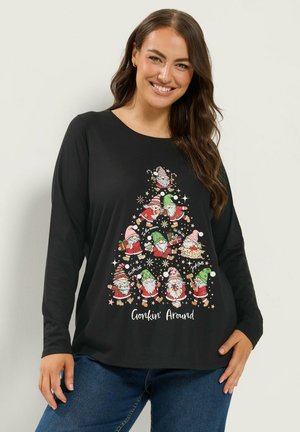 Femme aux longs cheveux bruns sourit, portant un t-shirt noir à manches longues avec un gnome de Noël, un sapin et le texte "Gonkin' Around".