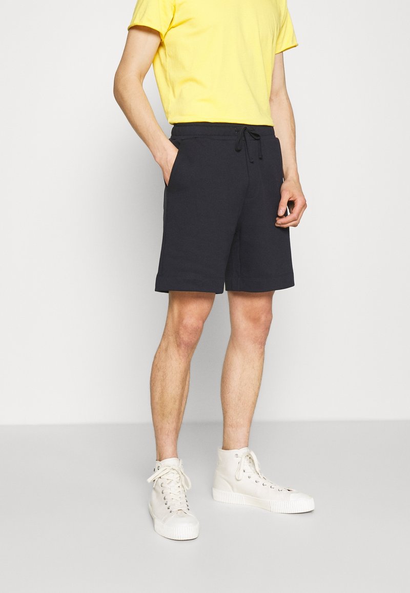 Shorts bleu marine en tissu mélangé coton avec une taille élastique et un cordon de serrage. Associés à un t-shirt jaune et des baskets blanches montantes.