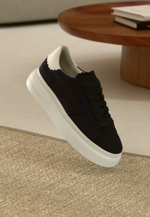 Czarny zamszowy sneaker z białym skórzanym tyłem, teksturowaną podeszwą i czarnymi sznurowadłami. Eleganckie, fazowane wykończenie na górnej części buta.