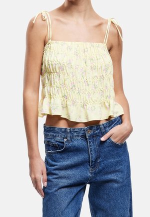 Kvinde iført en gul blomsterpræget smockcrop top med bindestraps kombineret med højtaljede blå jeans, den ene hånd i lommen.
