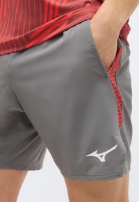 Άτομο που φορά γκρι αθλητικές βερμούδες με λευκό λογότυπο, κόκκινο και μαύρο ριγωτό πουκάμισο, με το χέρι στην τσέπη της βερμούδας.