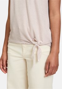 Haut léger à motifs en rose pâle avec un nœud à l'ourlet. Associé à un pantalon droit couleur crème. Détail de tissu texturé visible.