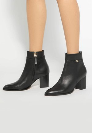 Bottines en cuir noir à talons blocs portées sur jambes nues, avec fermeture éclair latérale et petit détail de boucle dorée.