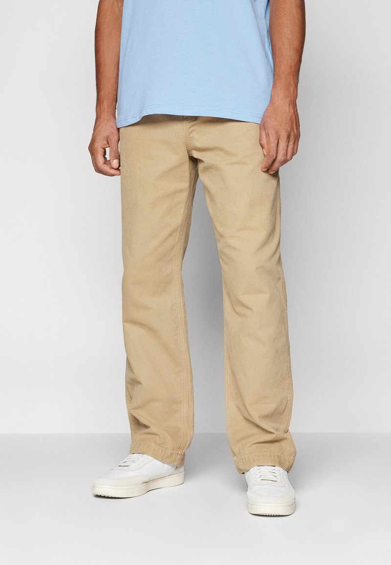 Beige cotton twill pants with a relaxed fit, straight legs, and side pockets, paired with white sneakers and a blue T-shirt.