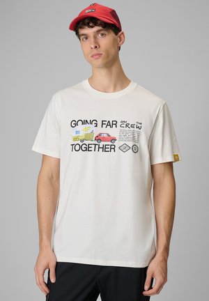 Młody mężczyzna ubrany w białą koszulkę z napisem „Going Far Together”, grafiką samochodu i kampera, oraz czerwoną czapkę z daszkiem, stoi na szarym tle.