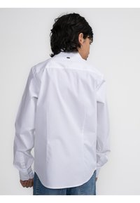 Witte lange mouw shirt van zachte stof, met knoopmanchetten en een afgeronde zoom. Heeft een klein zwart detail aan de achterkant van de nek.
