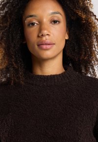 Brauner Strickpullover mit einer strukturierten, fuffy Oberfläche und einem gerippten Rundhalsausschnitt, getragen von einer Person mit lockigem Haar.