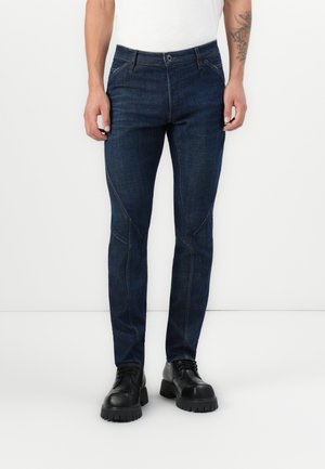 Mørkeblå denimjeans med slim fit, som har karakteristisk sømdetaljering og knappelukking, kombinert med svarte snørebåndsko.