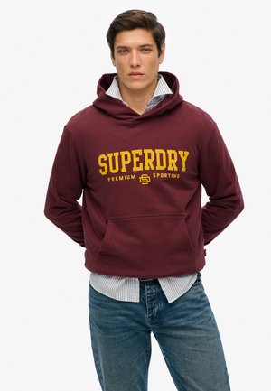 Sudadera con capucha granate con el logo amarillo "SUPERDRY", bolsillo delantero y capucha con cordón, superpuesta sobre una camisa de rayas azul claro y blanca.