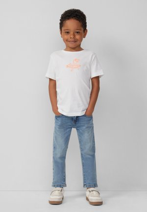 Witte t-shirt met korte mouwen en een oranje grafisch ontwerp, in combinatie met lichtblauwe jeans en beige sneakers, staand tegen een grijze achtergrond.