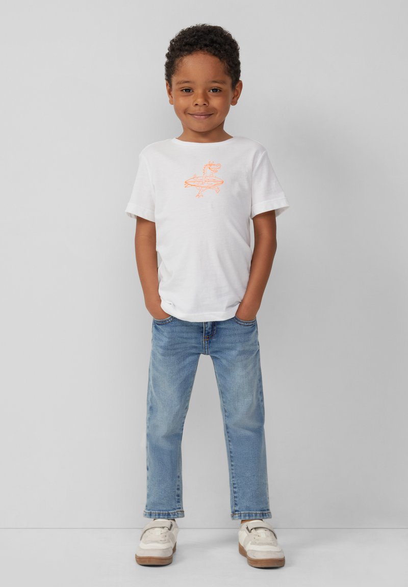 Witte t-shirt met korte mouwen en een oranje grafisch ontwerp, in combinatie met lichtblauwe jeans en beige sneakers, staand tegen een grijze achtergrond.
