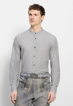 Jonge man draagt een grijs fijn gestreept overhemd met mandarijnkraag en knopen en grijze geborduurde traditionele leren broek met een decoratieve riem.