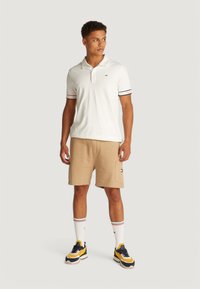 Camisa polo blanca con rayas rojas y azules en las mangas, pantalones cortos beige, calcetas blancas con acentos rojos y azules, y zapatillas negras con amarillo.