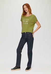 Olijfgroene t-shirt met witte tekst "LEEF IN HET NU," gecombineerd met donkerblauwe jeans en zwarte sneakers met een lichte zool.