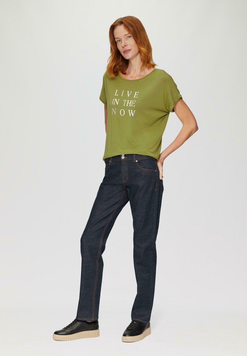 Olivengrünes T-Shirt mit weißem Text "LIVE IN THE NOW", kombiniert mit dunkelblauen Jeans und schwarzen Sneakers mit heller Sohle.