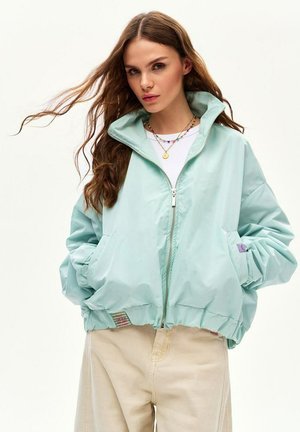 Kobieta z długimi brązowymi włosami, ubrana w jasnoniebieską kurtkę na zamek i beżowe spodnie, stojąca z rękami w kieszeniach na białym tle.