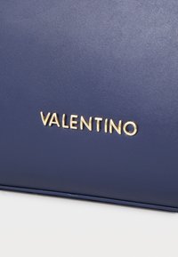 Ναυτικές δερμάτινες τσάντες με λεία υφή και χρυσό λογότυπο "VALENTINO" αποτυπωμένο μπροστά. Μινιμαλιστικός σχεδιασμός με καθαρές γραμμές.