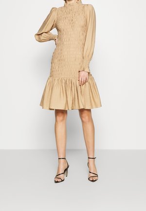 Vrouw in een beige lange mouw smock jurk met een gerimpelde zoom en zwarte bandjeshakken, staand tegen een witte muur.