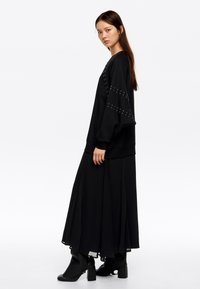 Sudadera negra de gran tamaño con detalles de tachuelas en las mangas, combinada con una falda larga negra y botas de tobillo con tacón cuadrado.