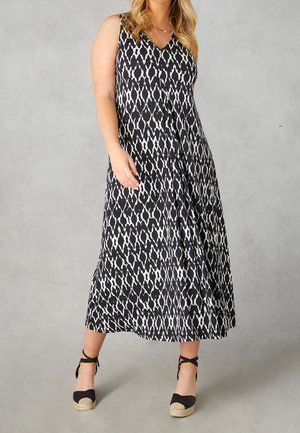 Nainen, joka käyttää hihattoman mustavalkoisen kuvioidun maximekon ja mustia nilkkaremmeillä varustettuja espadrille-pohjaisia kiilakorkoja, seisoo harmaata taustaa vasten.