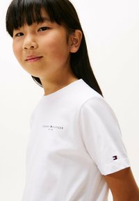 Ungdom med langt mørkt hår iført en hvit Tommy Hilfiger t-skjorte med logoer på brystet og ermet mot en ensfarget bakgrunn.