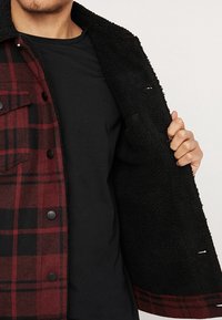 Rød og sort ternet jakke med sort, blødt, luddigt indre. Har knaplukning og brystlomme samt en blød, tekstureret foring.