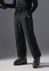 Pantalones de esquí negros con un cinturón, con acentos laterales en color amarillo neón, de corte holgado y cremalleras en el tobillo. Combinados con botas de esquí negras brillantes.
