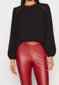 Femme portant un chemisier noir à manches longues avec des détails en dentelle et un pantalon taille haute en simili cuir rouge brillant.
