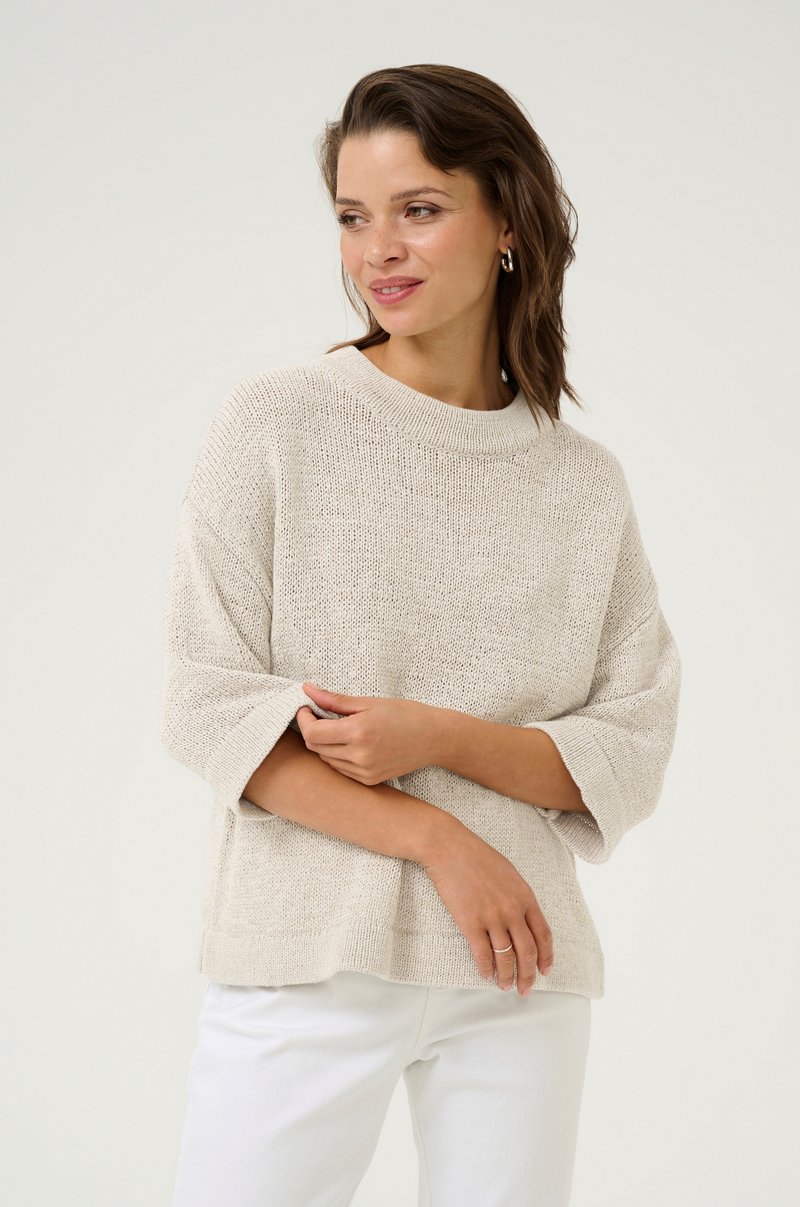 Pull en maille beige clair avec un large col et des manches trois-quarts, offrant une coupe décontractée et une texture aérée. Porté par-dessus un pantalon blanc.