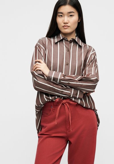 Νεαρή γυναίκα με μακριά μαύρα μαλλιά, που φοράει καφέ και λευκό πουκάμισο με κάθετες ρίγες και κόκκινο παντελόνι ψηλόμεσο με ζώνη δεμένη στη μέση.