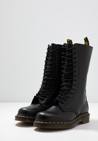 Bottes de combat en cuir noir à lacets avec coutures jaunes et languettes, placées sur une surface blanche devant un fond neutre.
