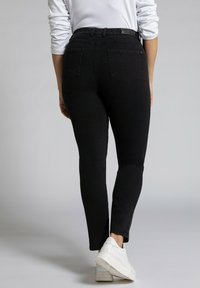 Musta korkeavyötäröinen slim fit -farkut, joissa on viisi taskua ja sileä pinta, yhdistettynä valkoisiin tennareihin neutraalissa taustassa.