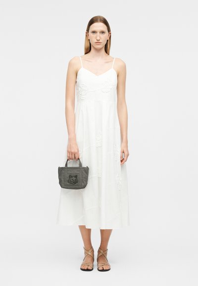 Γυναίκα με λευκό μίντι φόρεμα χωρίς μανίκια, κρατώντας μια μικρή γκρι τσάντα χειρός με ζωικό μοτίβο, φορώντας μπεζ σανδάλια με λουράκια, στέκεται μπροστά σε απλό φόντο.