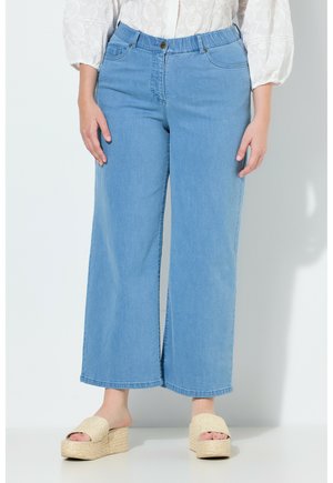 Frau trägt hellblaue Jeans mit weitem Bein, eine weiße Bluse mit strukturierten Mustern und beige Plateausandalen aus gewebtem Material.