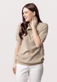 Beige gebreide polo trui met korte mouwen, voorzien van een kraag en een sluiting met drie knopen, gedragen met witte broeken.