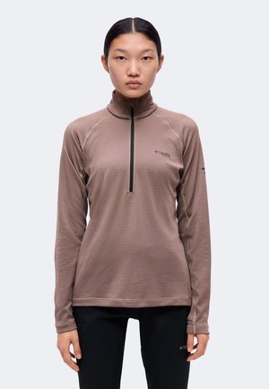 Femme aux cheveux foncés en chignon bas porte un haut marron à manches longues avec fermeture éclair et un pantalon noir, debout face à un fond uni.