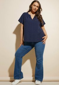 Top blu navy con scollo a V e maniche corte, caratterizzato da un motivo testurizzato, abbinato a jeans chiari a campana e sneakers bianche.