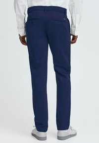 Tumši zilā krāsā taisnā griezuma bikses, ar divām aizmugurējām kabatām ar pogām un kontrastējošu diegu. Valkā ar gaiši pelēkiem sporta apaviem.