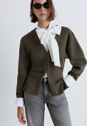 Frau mit schwarzen Sonnenbrillen, weißer Bluse mit großer Schleife, olivgrünem Cardigan und grauen Jeans, Hand in der Hüfte vor schlichter Hintergrund.