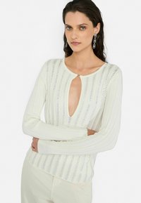 Maglione bianco a coste con scollo a V profondo e un accento argentato. La texture presenta righe verticali di piccoli dettagli lucidi.