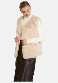 Gilet in pelliccia sintetica beige con una superficie morbida, design aperto sul davanti e senza maniche, abbinato a un dolcevita color crema e una gonna di pelle scura.