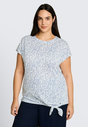 Frau mit langen dunklen Haaren, die ein weißes Kurzarmtop mit blauem Blumenmuster und Frontknoten trägt, kombiniert mit dunkler Hose.